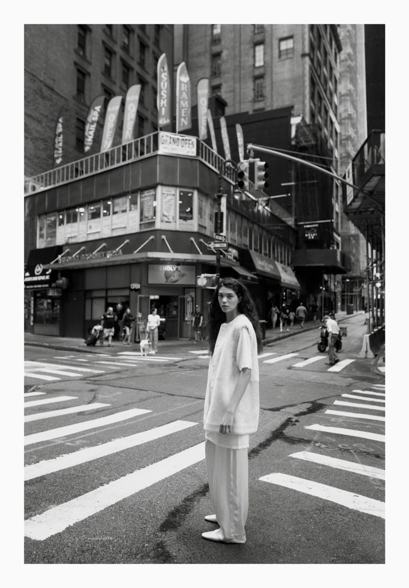 Monochrome full shot of a woman in white outfit standing on a crosswalk in a city, with tall buildings and people in the background.