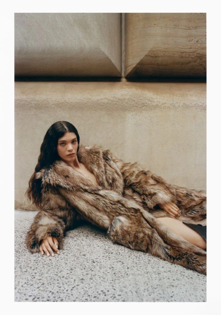 Brunette woman in a fur coat poses indoors against a textured wall. Fashionable outerwear with long dark hair and a serious expression.