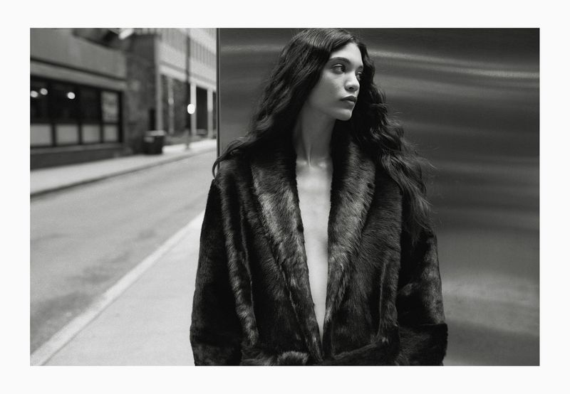Monochrome portrait of a young woman with long wavy hair wearing a fur coat, looking away, in an urban environment, capturing a fashion editorial vibe.