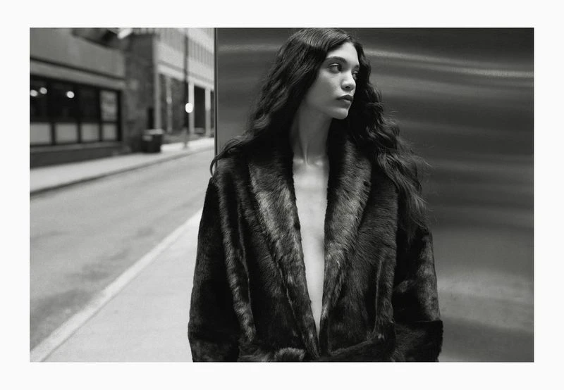 Monochrome portrait of a young woman with long wavy hair wearing a fur coat, looking away, in an urban environment, capturing a fashion editorial vibe.