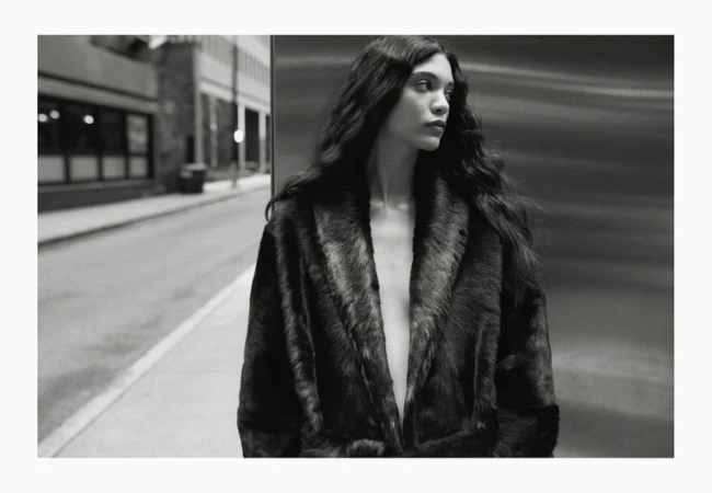 Monochrome portrait of a young woman with long wavy hair wearing a fur coat, looking away, in an urban environment, capturing a fashion editorial vibe.