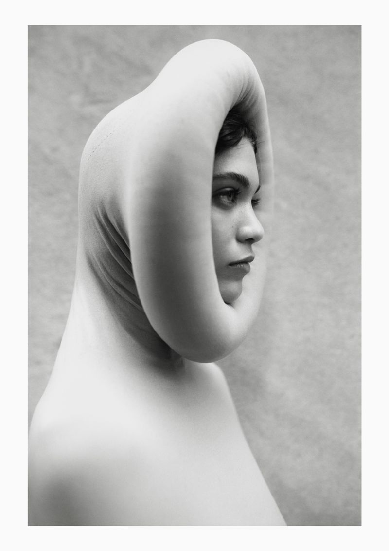 Monochrome portrait of a woman in a sculptural head covering, gazing pensively with a smooth skin tone and neutral expression against a textured backdrop.