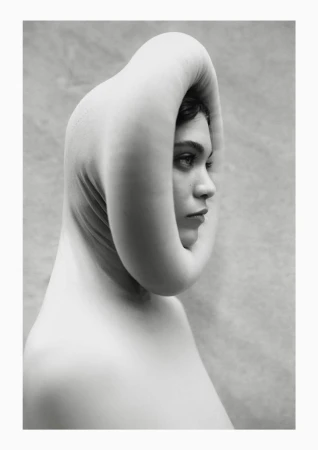 Monochrome portrait of a woman in a sculptural head covering, gazing pensively with a smooth skin tone and neutral expression against a textured backdrop.