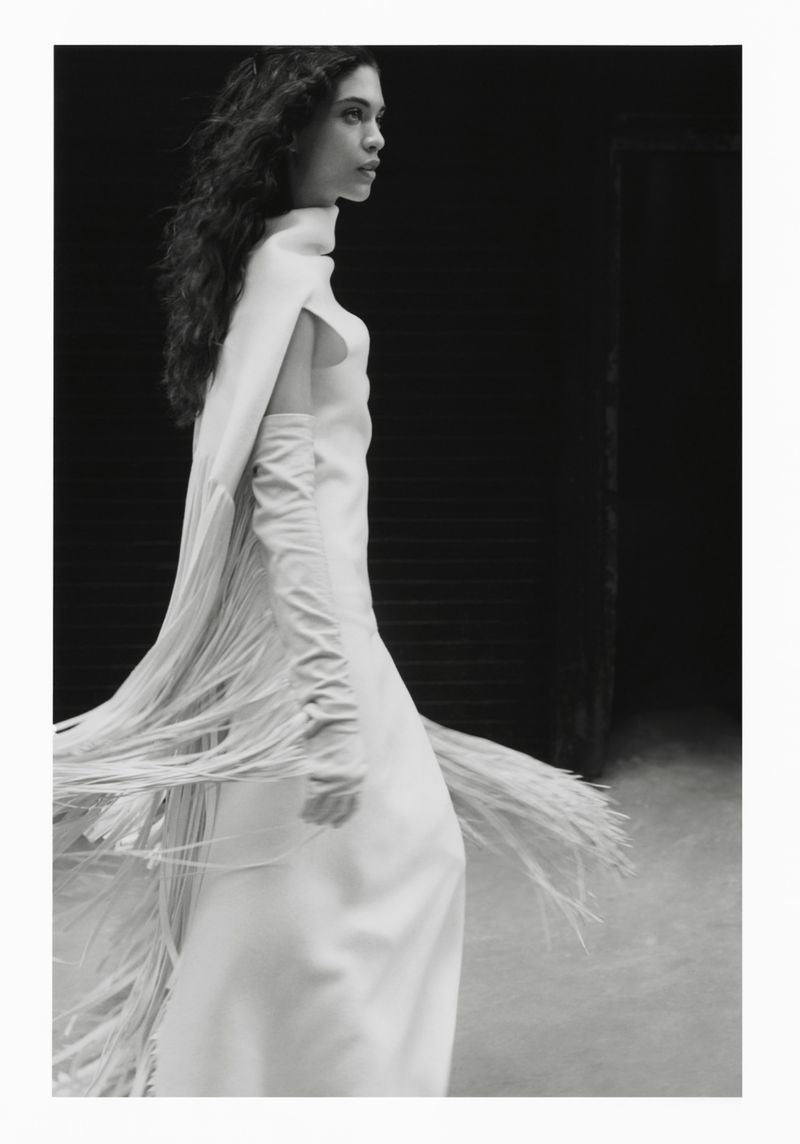 Monochrome image of a woman in a white gown with fringe detailing, captured in a low-light setting, creating a timeless, elegant aesthetic.