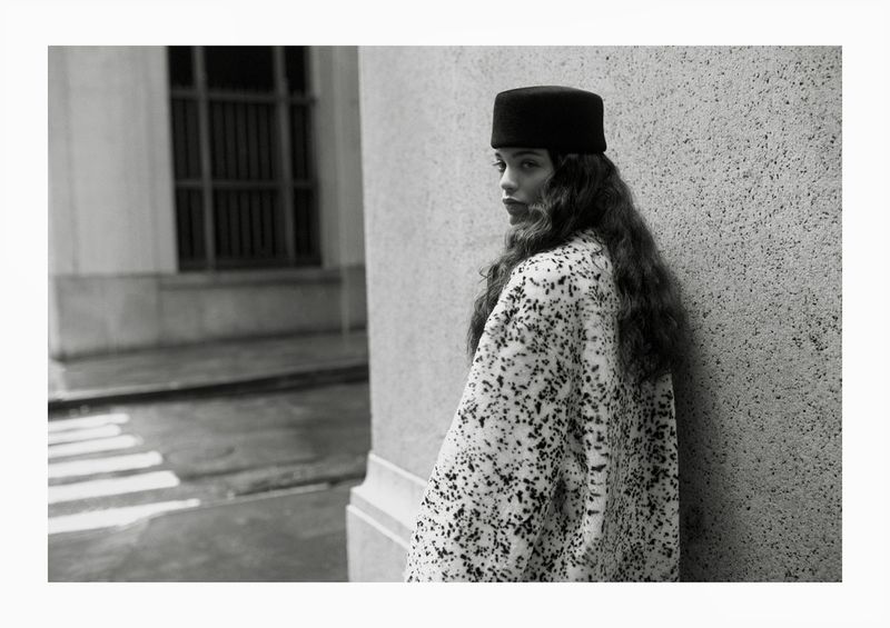 Black and white portrait of a woman with long, wavy hair wearing a hat and patterned coat, posing in front of a concrete wall.