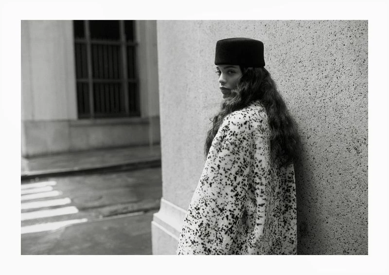 Black and white portrait of a woman with long, wavy hair wearing a hat and patterned coat, posing in front of a concrete wall.