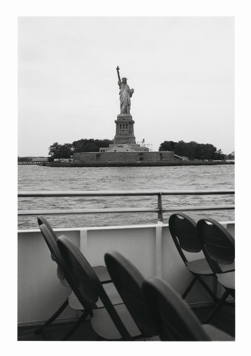 Black and white outdoor shot of the Statue of Liberty on Liberty Island, viewed from a boat, with rows of folding chairs in foreground.