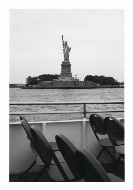 Black and white outdoor shot of the Statue of Liberty on Liberty Island, viewed from a boat, with rows of folding chairs in foreground.
