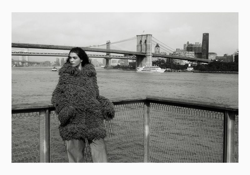A woman in a textured coat stands near a railing with the Brooklyn Bridge and the New York City skyline in the background on a cloudy day.