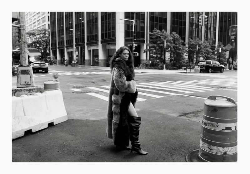 A woman in a fur coat and high boots is standing on a city street in black and white. Tall buildings visible behind her.