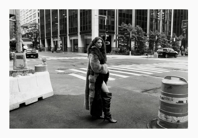 A woman in a fur coat and high boots is standing on a city street in black and white. Tall buildings visible behind her.