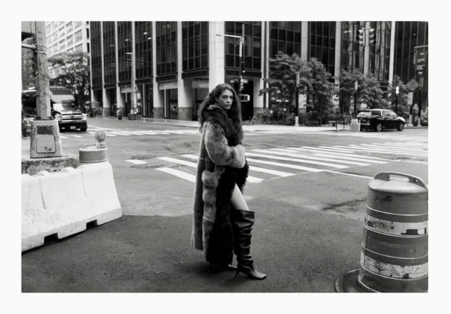 A woman in a fur coat and high boots is standing on a city street in black and white. Tall buildings visible behind her.