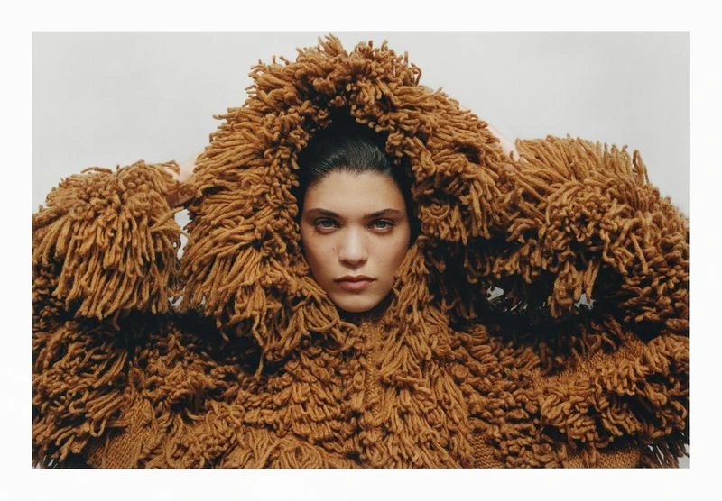 Portrait of a woman in a brown, shaggy, textured sweater with a hood. The background is a neutral grey in a studio shot.