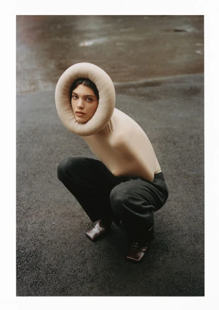 Fashionable woman in a beige bodysuit and dark pants poses while squatting on asphalt, wearing a padded ring around her head.