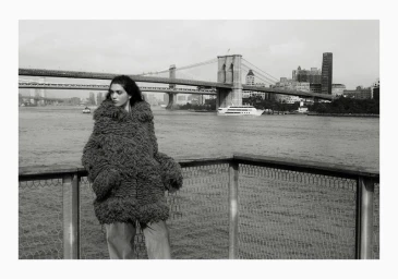 Black and white photo of model Marina Moioli in a shaggy oversized coat standing by a waterfront railing before the Brooklyn Bridge.