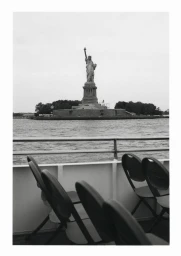 Black and white view of the Statue of Liberty across the water framed from the deck of a ferry with empty passenger seats.