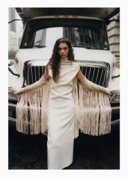 Model Marina Moioli poses in a floor-length cream high-neck dress with long fringed sleeves in front of a white commercial truck.