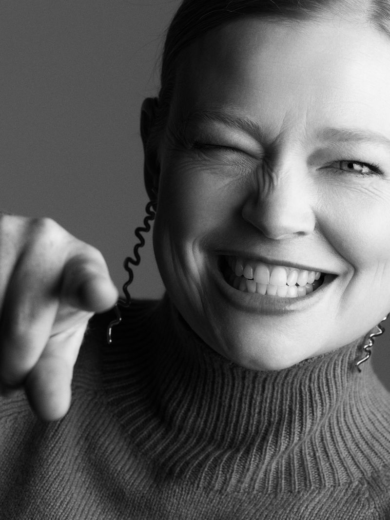 Monochrome portrait of a smiling adult woman pointing with one eye closed and wearing spiral earrings against a grey backdrop.