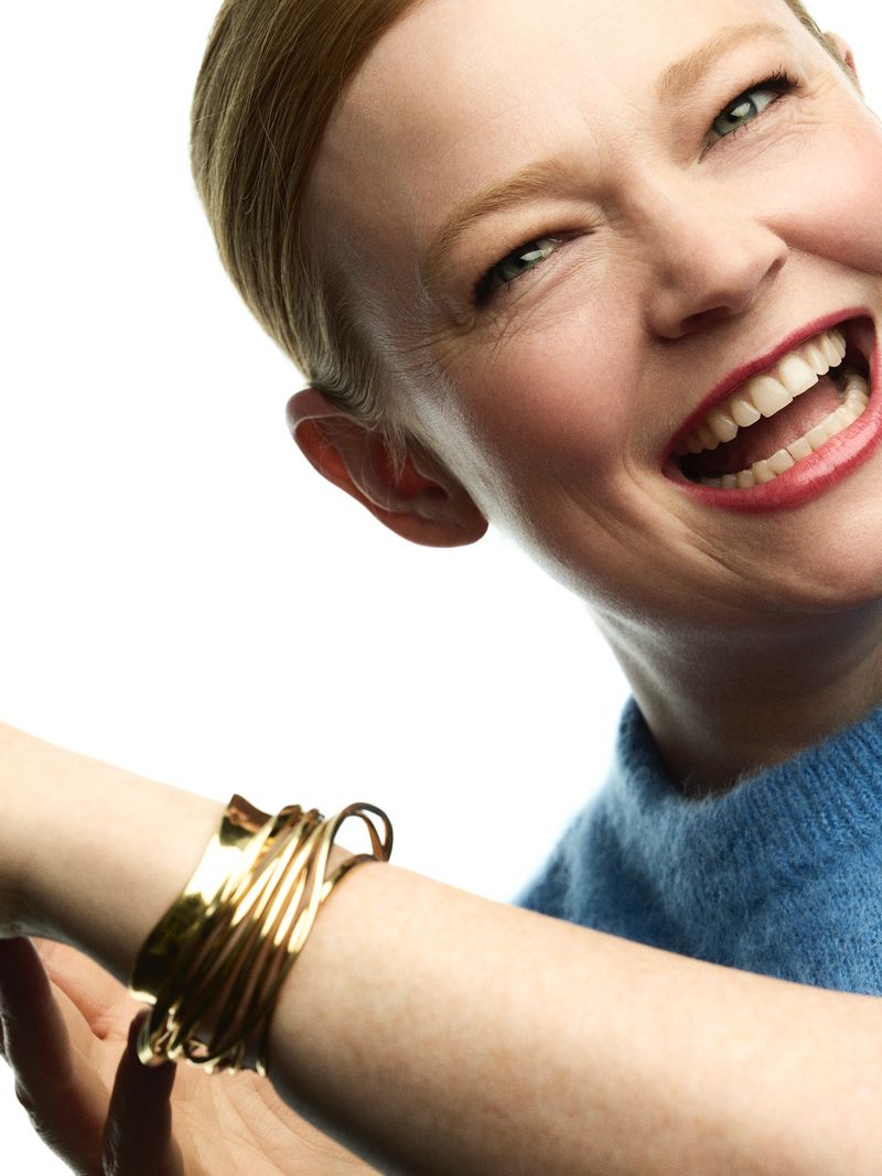Close-up of an adult female smiling, wearing a blue sweater and a gold bracelet against a white background.
