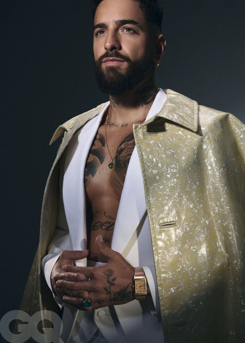 Portrait of a bearded adult male celebrity, Maluma, in a light yellow patterned coat, vest, and luxury watch against a grey background.
