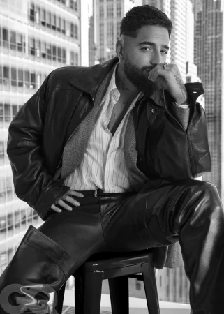 Black and white portrait of adult male seated in leather pants and jacket with cityscape background, GQ logo is visible in the foreground.