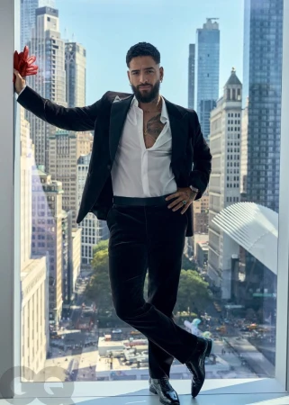 Maluma in a black suit and white shirt poses in front of a large window with a city view, adding a touch of elegance and celebrity charm.