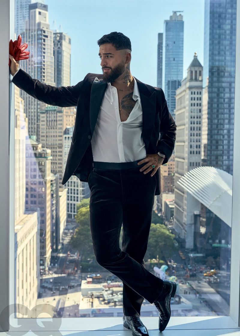 A man in a black suit and unbuttoned white shirt poses indoors next to a large window overlooking an urban cityscape.