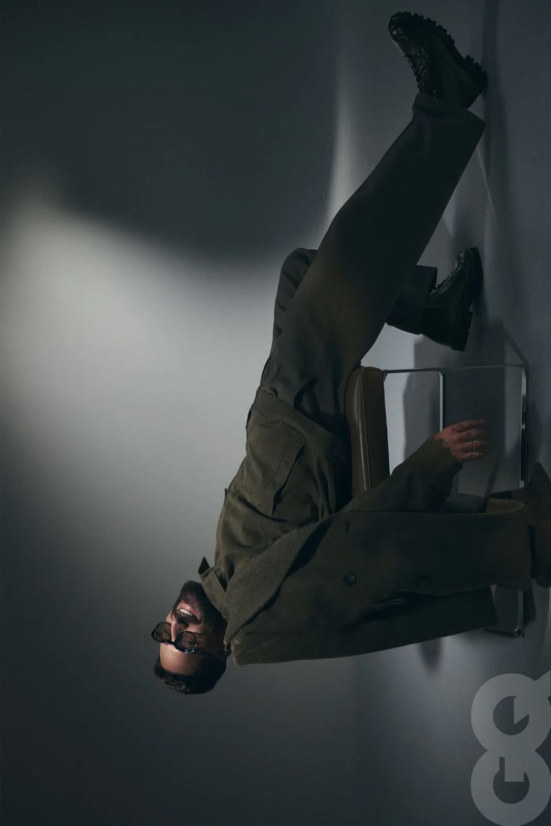 Man in olive green suit and sunglasses poses upside down on a chair against a gray wall.  GQ logo visible in corner.