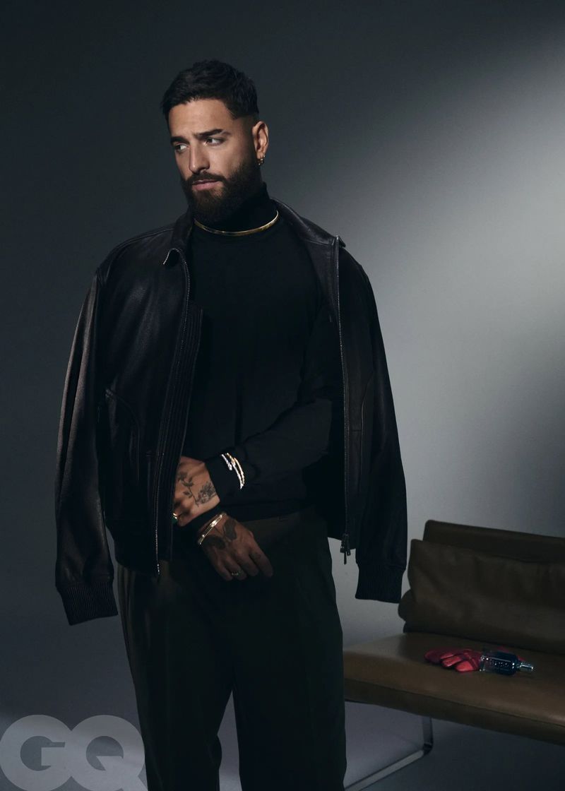 Portrait of a bearded man with a leather jacket draped over his shoulder, wearing a turtleneck, standing in a studio with a chair.