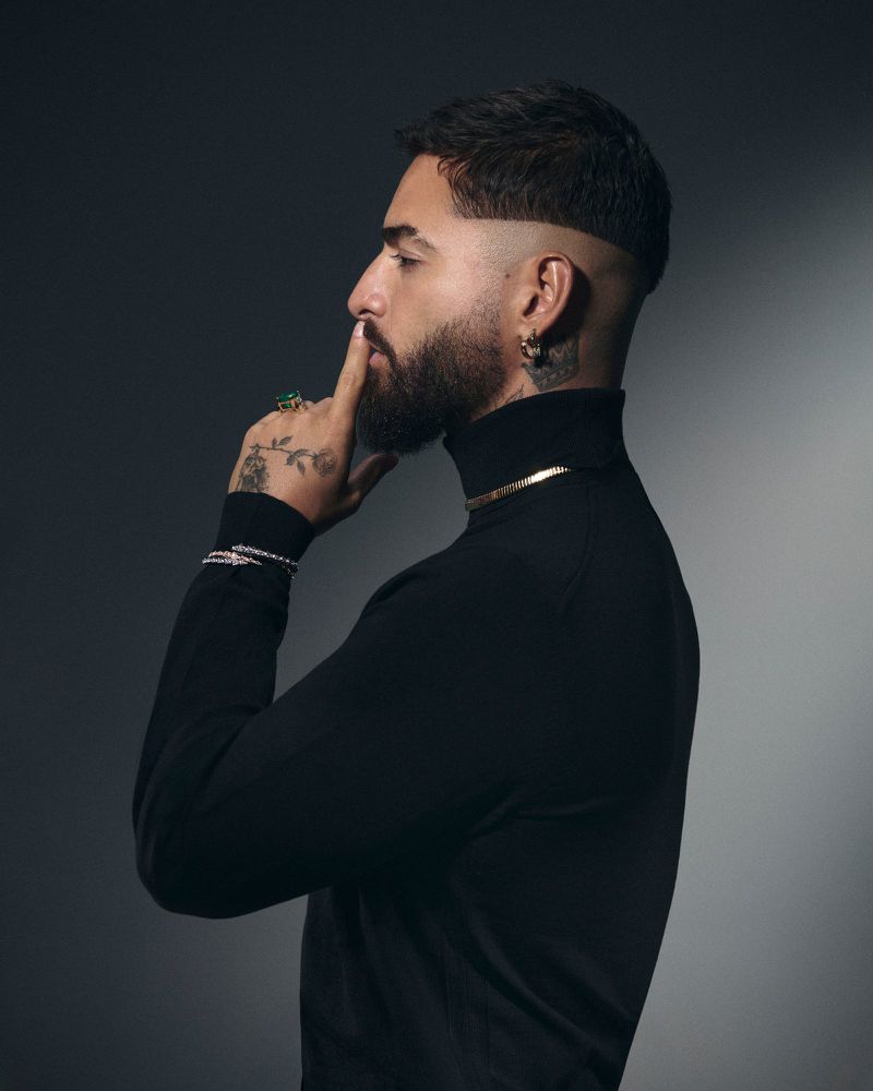 Portrait of an adult male with a beard wearing a black turtleneck and gold jewelry, holding a finger to his lips, against a gray backdrop.
