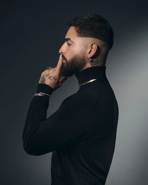 Portrait of an adult male with a beard wearing a black turtleneck and gold jewelry, holding a finger to his lips, against a gray backdrop.