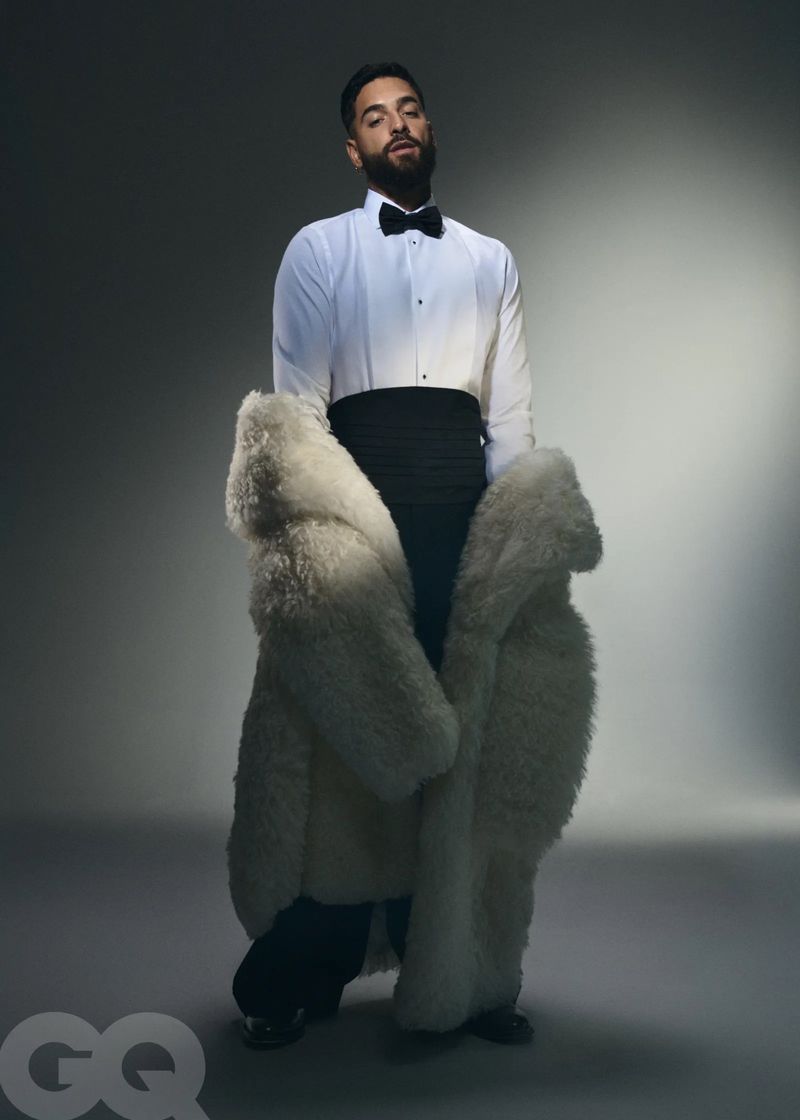A bearded man poses in formal wear and a large white fur coat in a studio setting with gray gradient background and dramatic lighting.