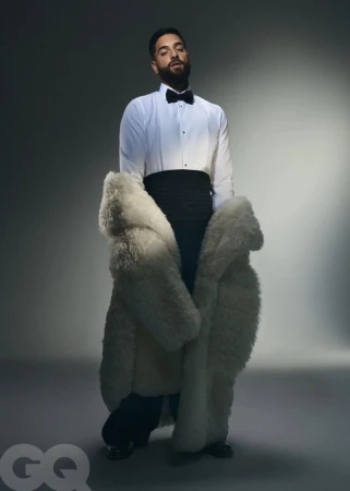 A bearded man poses in formal wear and a large white fur coat in a studio setting with gray gradient background and dramatic lighting.