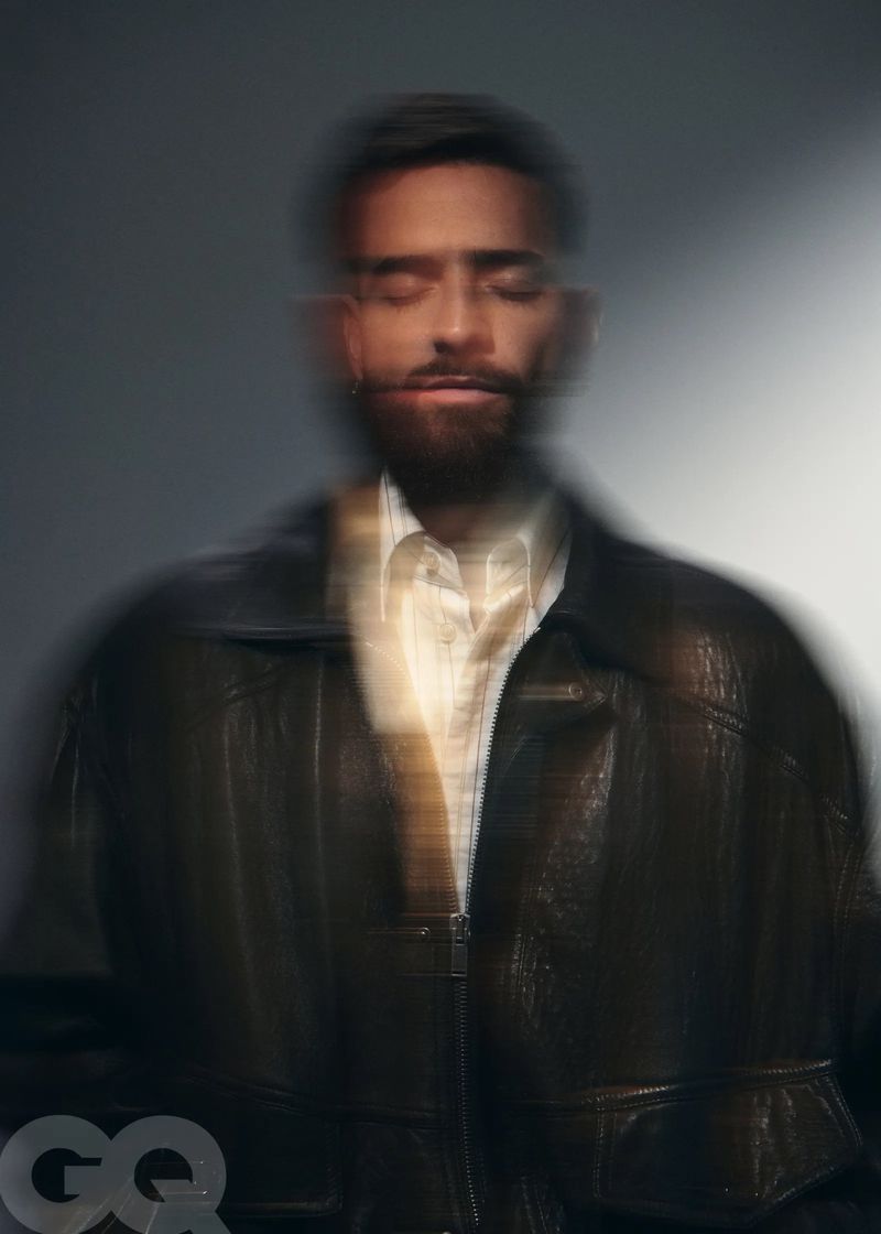 Portrait of a blurry man with a beard, wearing a jacket, with a light background and a GQ logo in the lower-left corner.