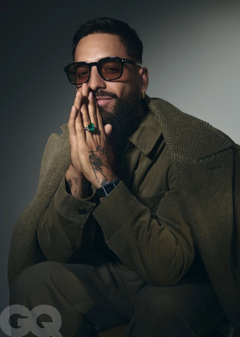 Portrait of a bearded adult male wearing sunglasses, an olive green jacket, and a large emerald ring, posing indoors against a grey background.