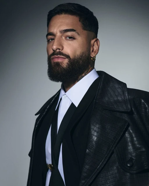 Close-up studio portrait of a man with a beard wearing a white shirt, black suit, and black leather trench coat, against a gray background.