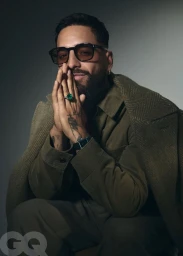 Maluma wearing tinted sunglasses and an olive green coat with clasped hands against a studio backdrop.
