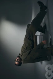 Maluma wearing an olive green suit and dark glasses, reclining on a chair against a spotlighted studio background for GQ Mexico.