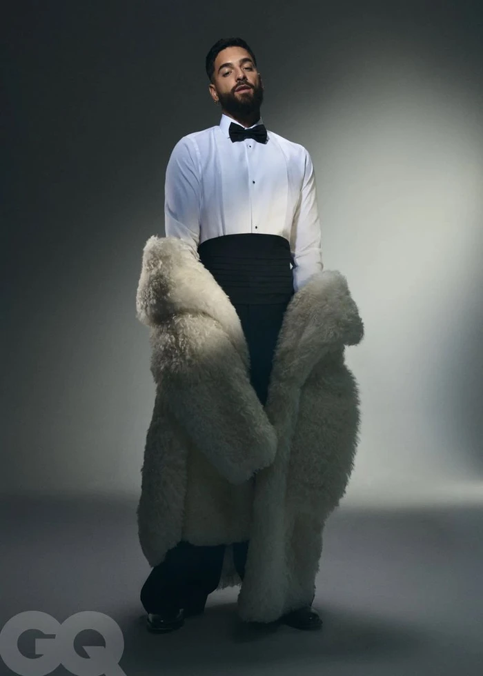 Maluma posing in a white tuxedo shirt with a black bow tie, cummerbund, and an oversized white fur coat against a grey backdrop.