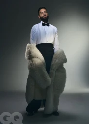 Maluma posing in a white tuxedo shirt with a black bow tie, cummerbund, and an oversized white fur coat against a grey backdrop.