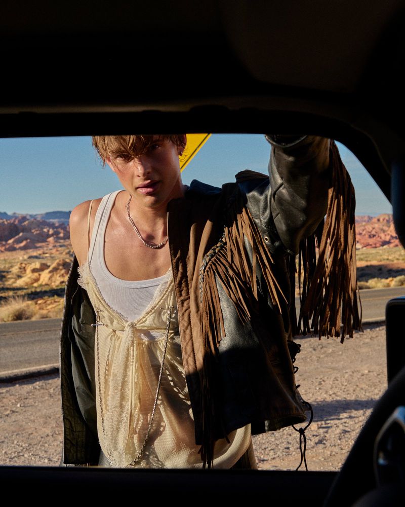 A young adult male stands by a car in a desert landscape, wearing a white tank top and jacket with fringe, under bright sunlight.