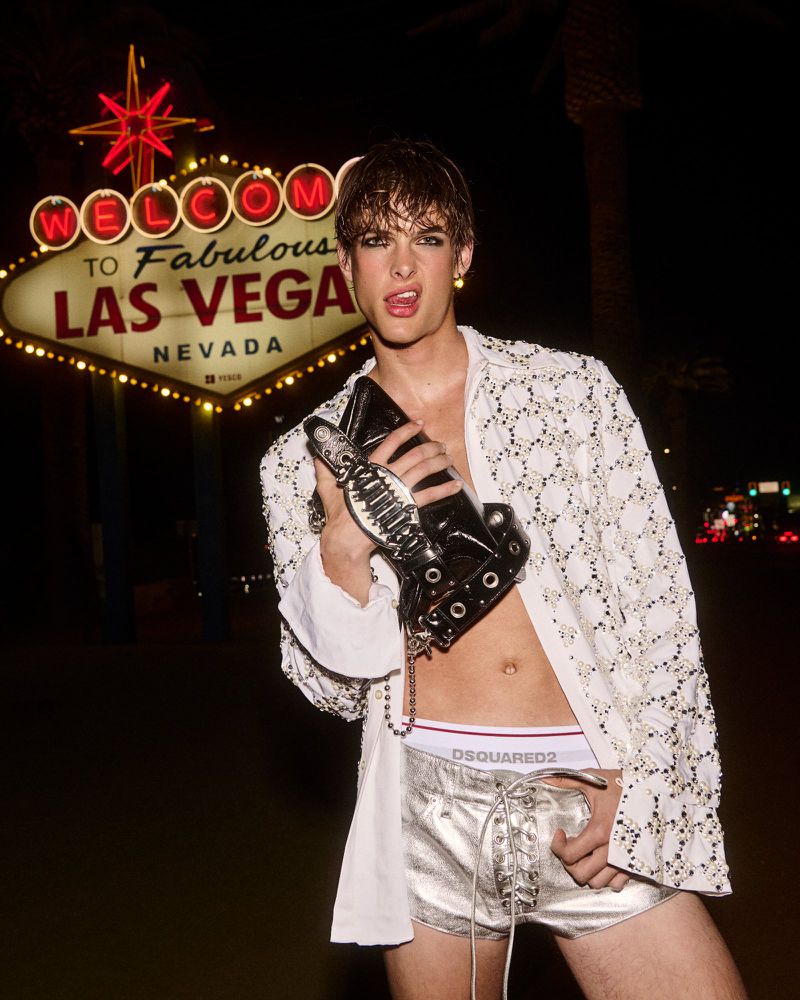 Man poses in Las Vegas wearing silver shorts, open white shirt, and black platform boots, with the iconic 'Welcome' sign in background.