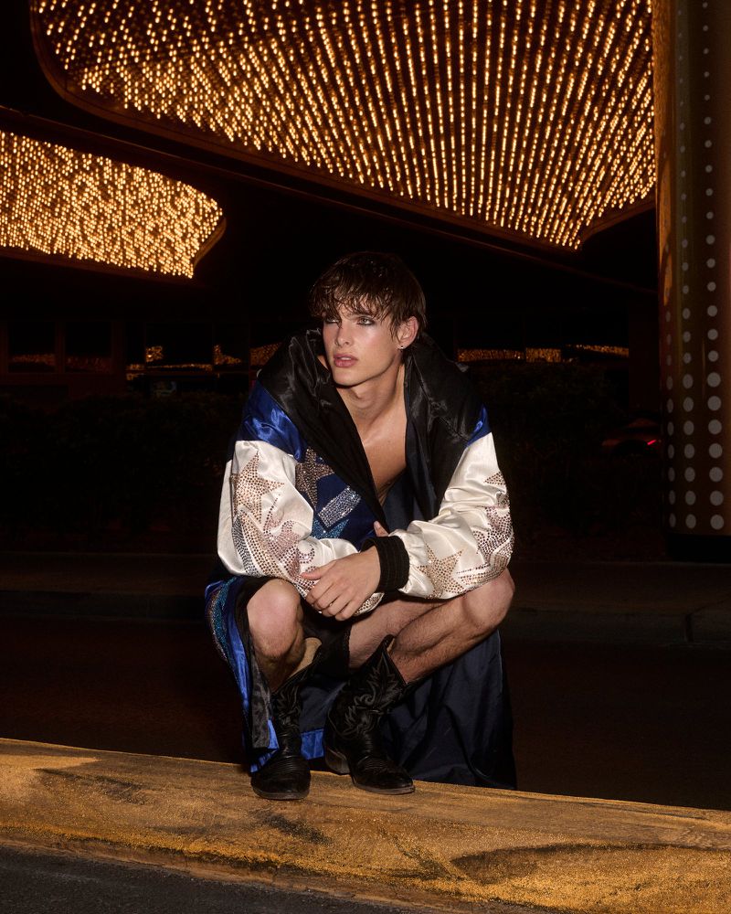 Young adult male model squats in a robe and boots outdoors under bright marquee lights at night for a fashion editorial shoot.