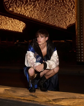 Young adult male model squats in a robe and boots outdoors under bright marquee lights at night for a fashion editorial shoot.