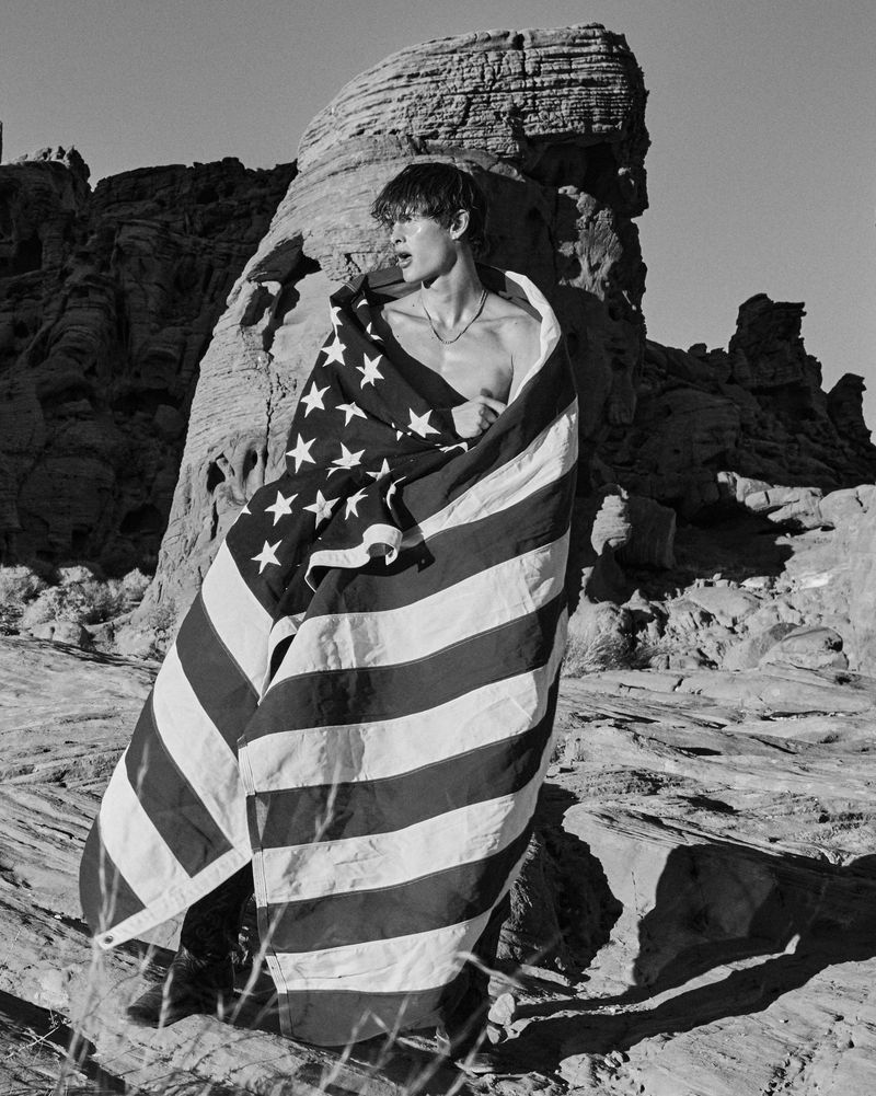 A young man stands wrapped in an American flag in a rocky, outdoor desert setting in black and white, under a clear sky.