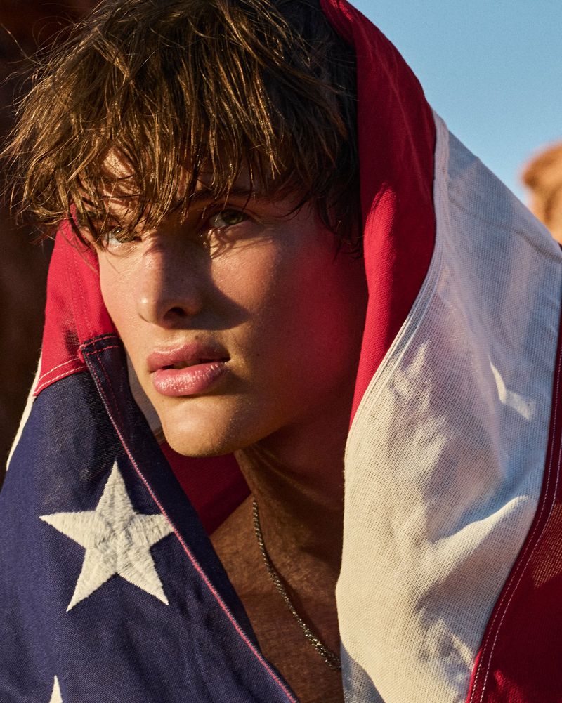 Young male model wrapped in an American flag with direct sunlight illuminating his face against a natural outdoor setting