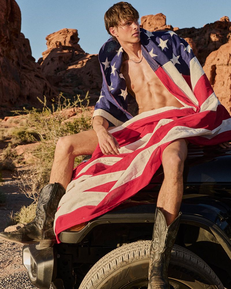Shirtless man sits on a car in a desert landscape draped in an American flag, wearing black cowboy boots against a blue sky.