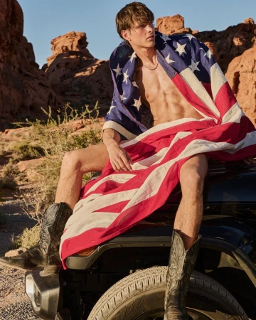 Shirtless man sits on a car in a desert landscape draped in an American flag, wearing black cowboy boots against a blue sky.