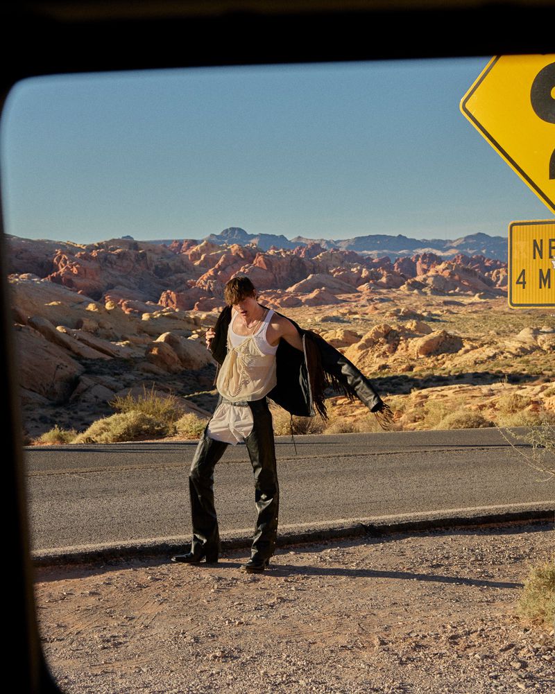 Model poses on a roadside in black chaps, a white top, and jacket against a backdrop of desert mountains and a clear blue sky.