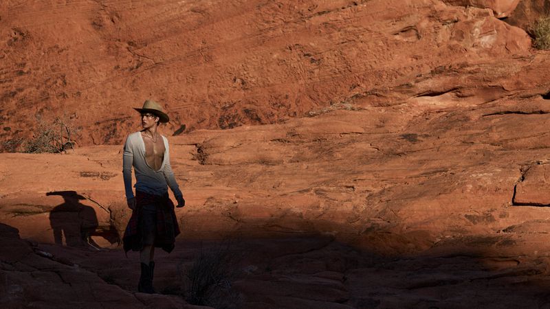 A person wearing a cowboy hat, long sleeve shirt, and skirt poses in a desert landscape with red rock formations in the background.